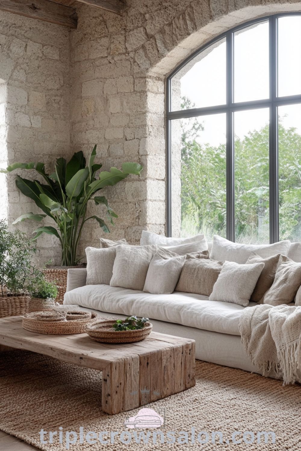 Cozy living room with earthy tones, textured stone walls, warm wooden floors, large windows, indoor plants, plush handwoven rug, rustic coffee table, cozy throws, and woven baskets showcasing nature-inspired decor ideas for your home. Visit triplecrownsalon.com for more unique ideas.